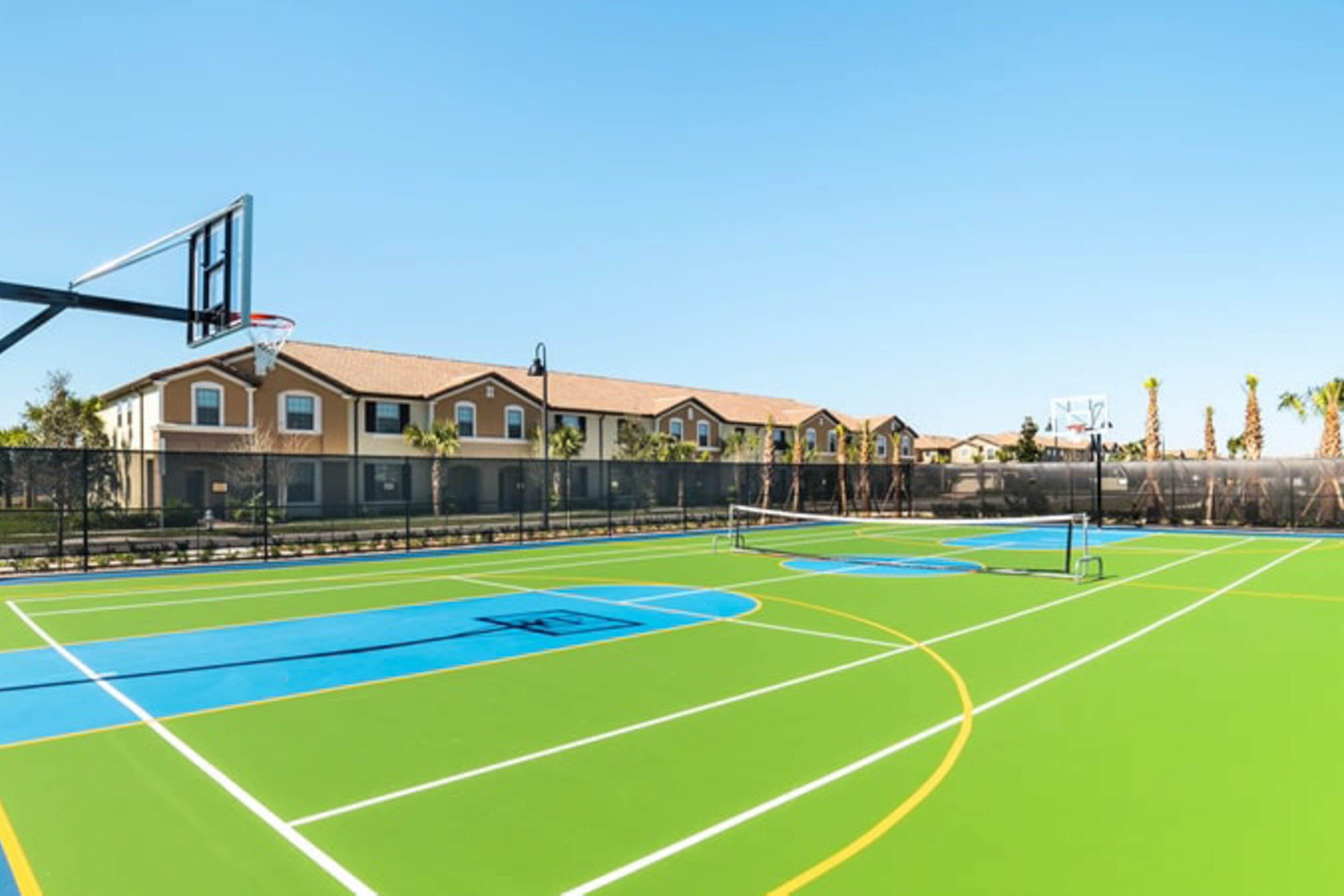 An outdoor basketball court with a clear sky, surrounded by a fence and a row of houses.