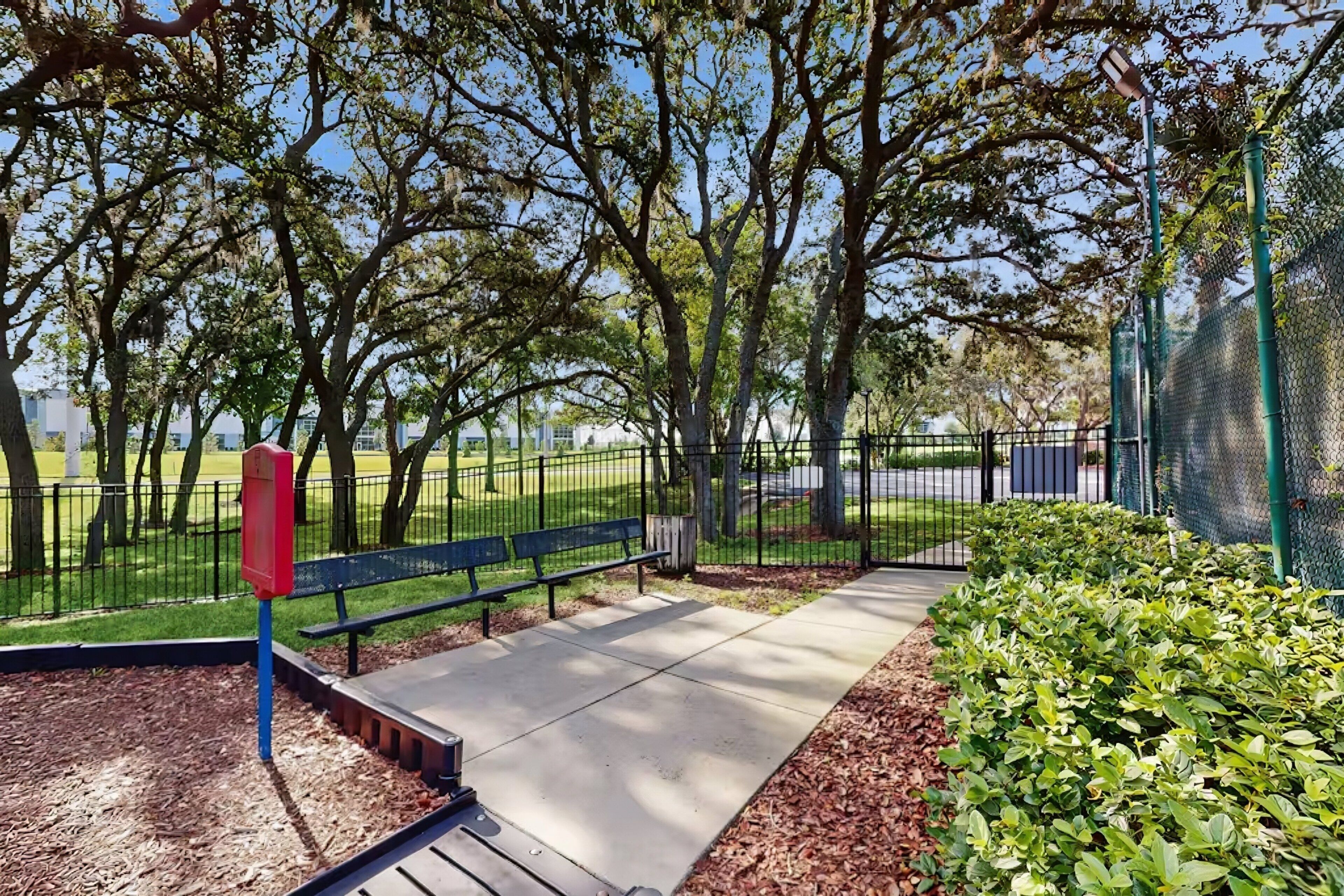 A park with a paved walkway, benches, a fence, and trees.
