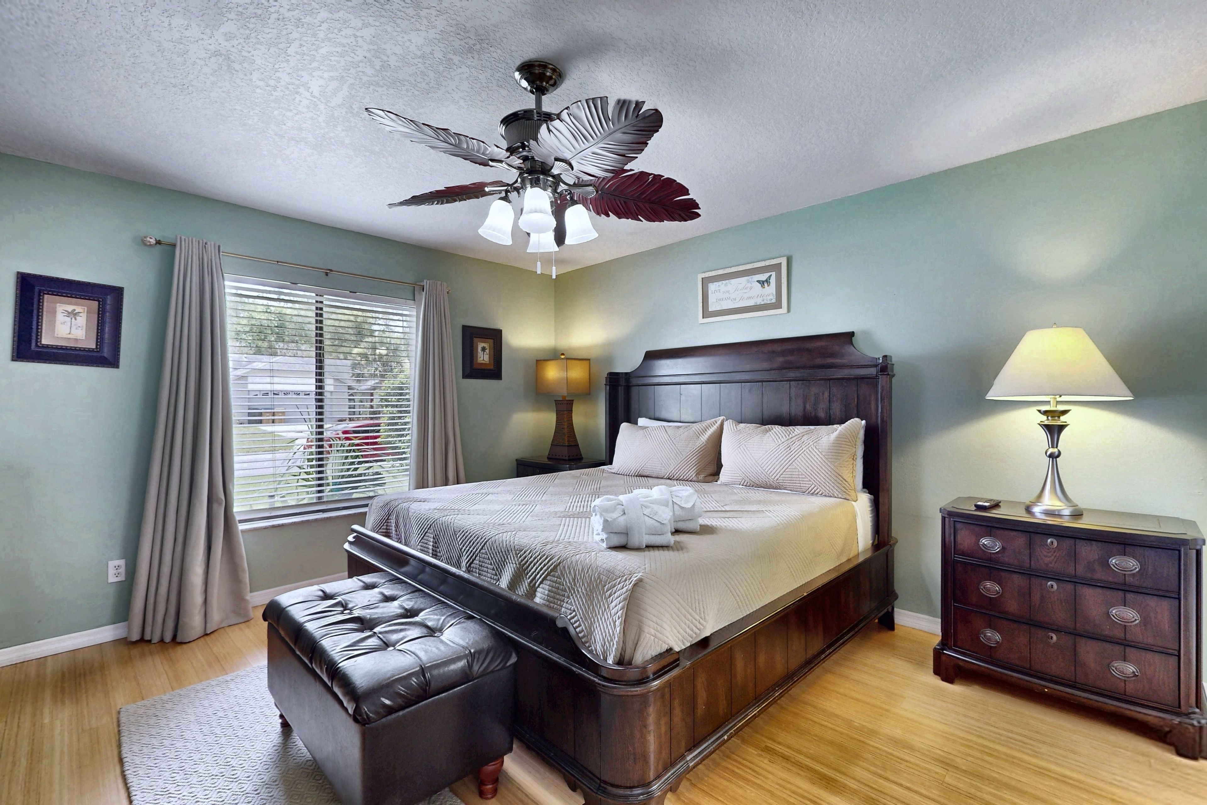 A bedroom with a large bed, a wooden nightstand, a lamp, a ceiling fan, a window with blinds, and a view of the outdoors.