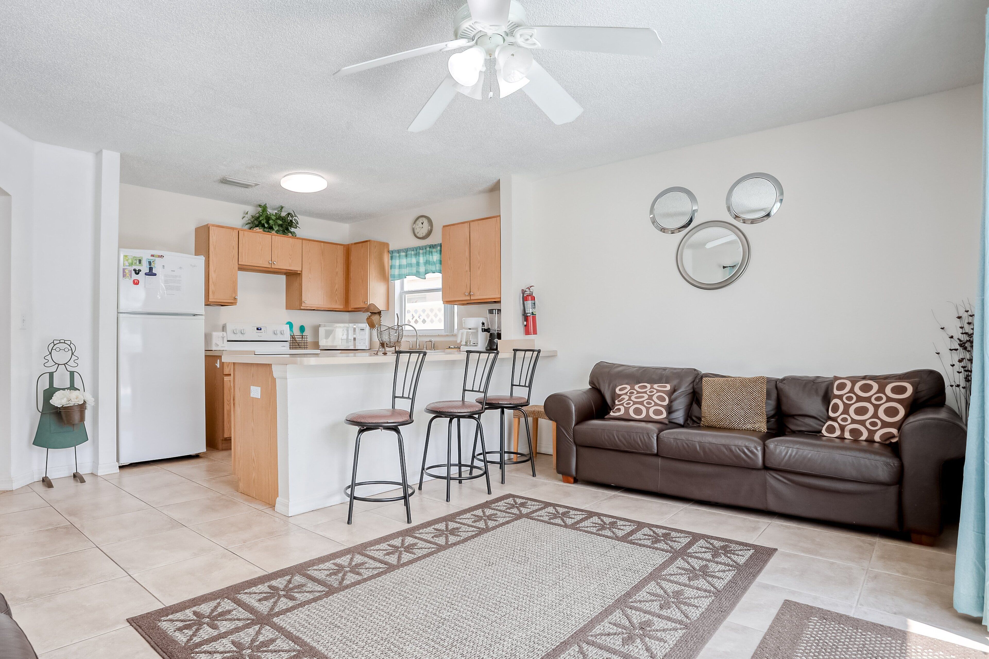 A modern kitchen with a central island, bar stools, and a dining area. There is a brown leather sofa with patterned cushions in the living room.