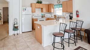 A modern kitchen with wooden cabinets, a white refrigerator, a microwave, a stove, and a coffee maker. There are four bar stools and a small island with a sink. - Enchanting Dog-friendly Home With Pool, Lanai, Central AC, & Washer/dryer (Kissimmee)