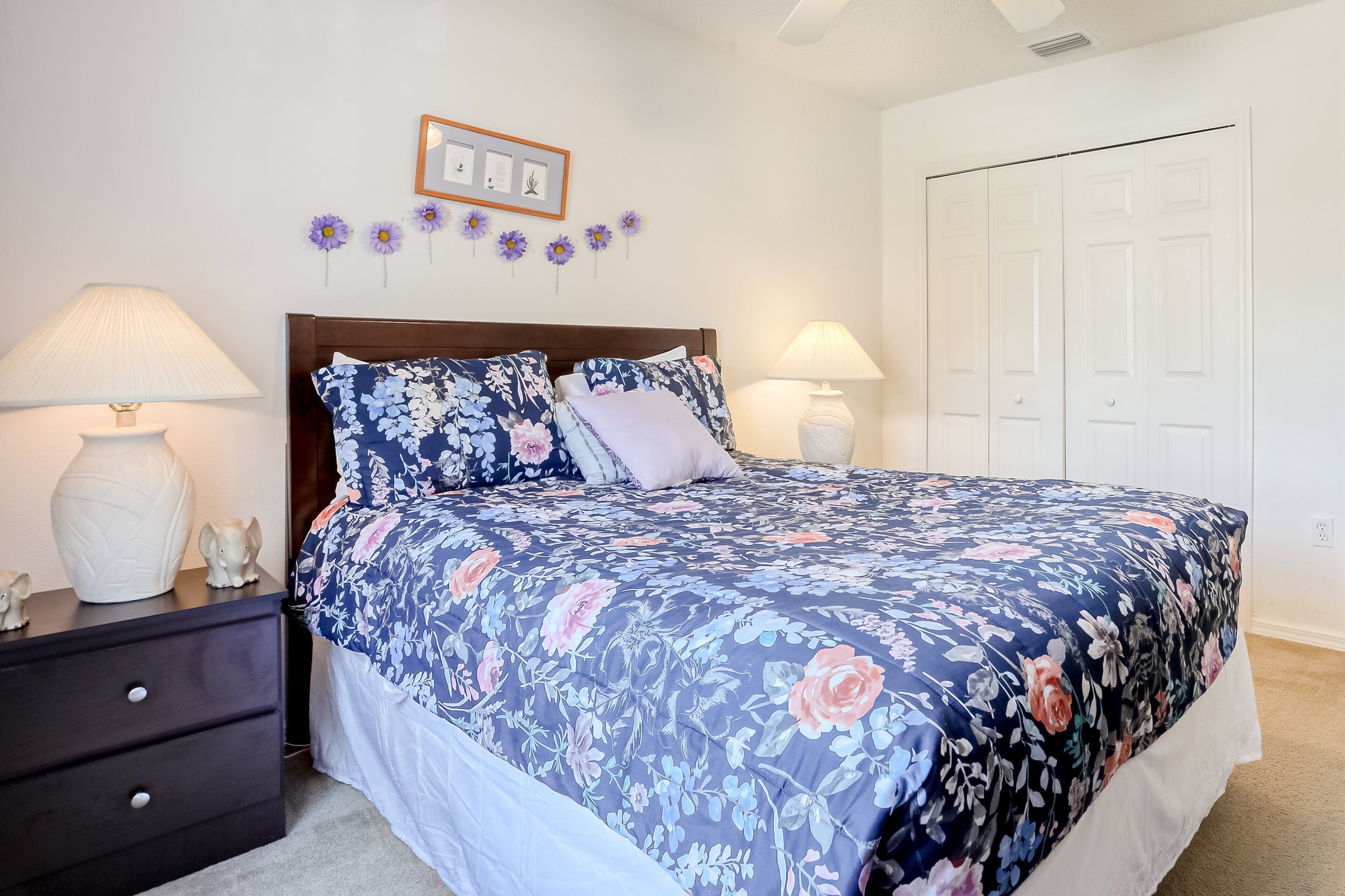 Un dormitorio con edredón floral, dos lámparas de noche, una mesita de noche de madera oscura, un jarrón blanco y un cuadro en la pared.