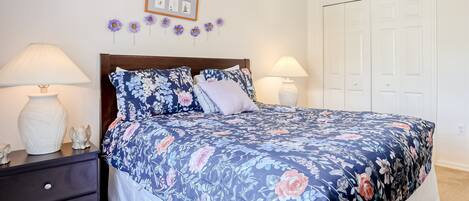 Un dormitorio con edredón floral, dos lámparas de noche, una mesita de noche de madera oscura, un jarrón blanco y un cuadro en la pared.