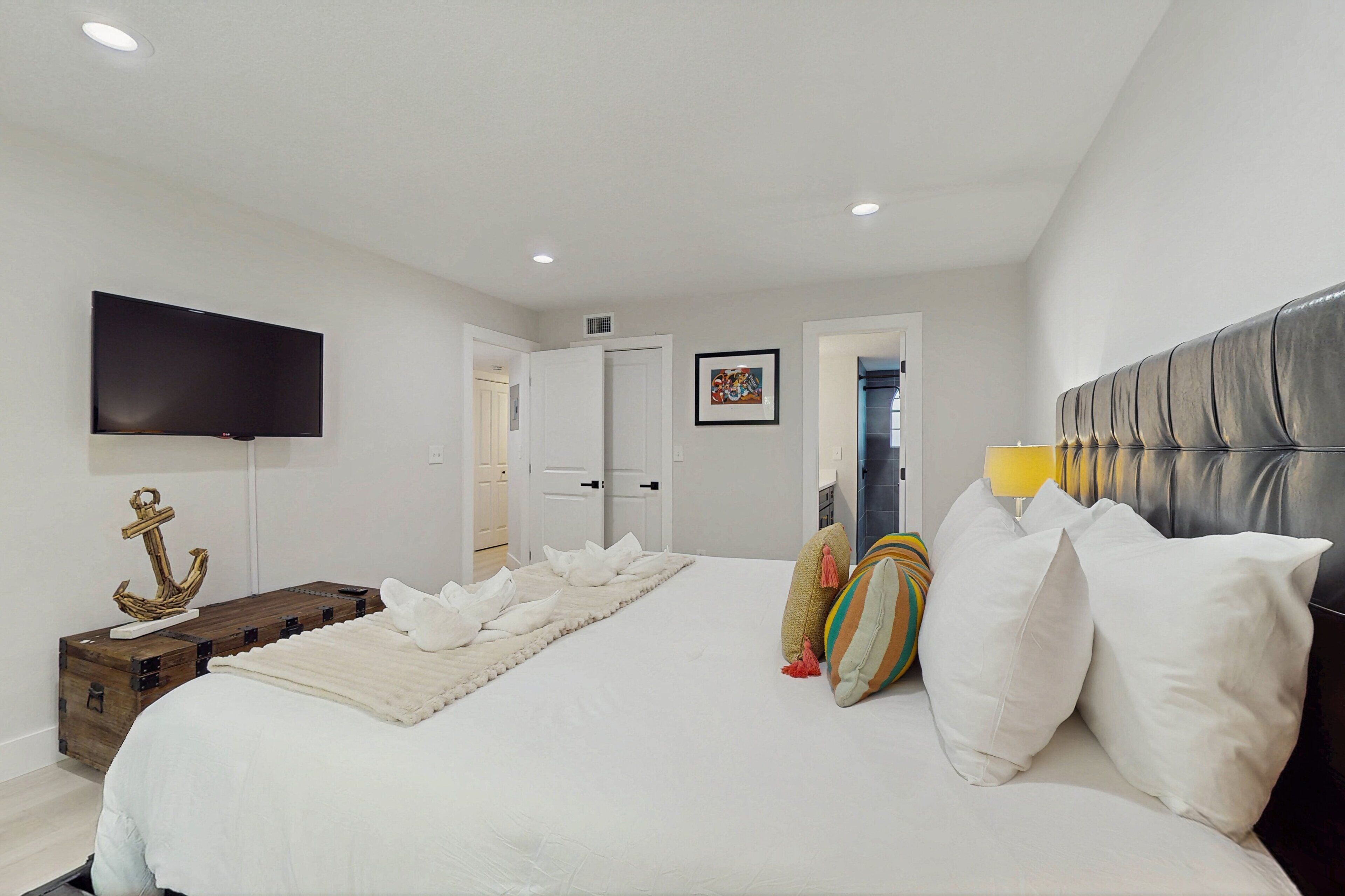 A bedroom with a large bed, a flat-screen TV, a wooden chest, and a door leading to another room.