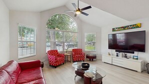 A spacious living room with a red sofa, two patterned armchairs, a glass coffee table, and a flat-screen TV on a white cabinet. - Beautiful Home Boasting Entertainment for Everyone With Wifi (Kissimmee)