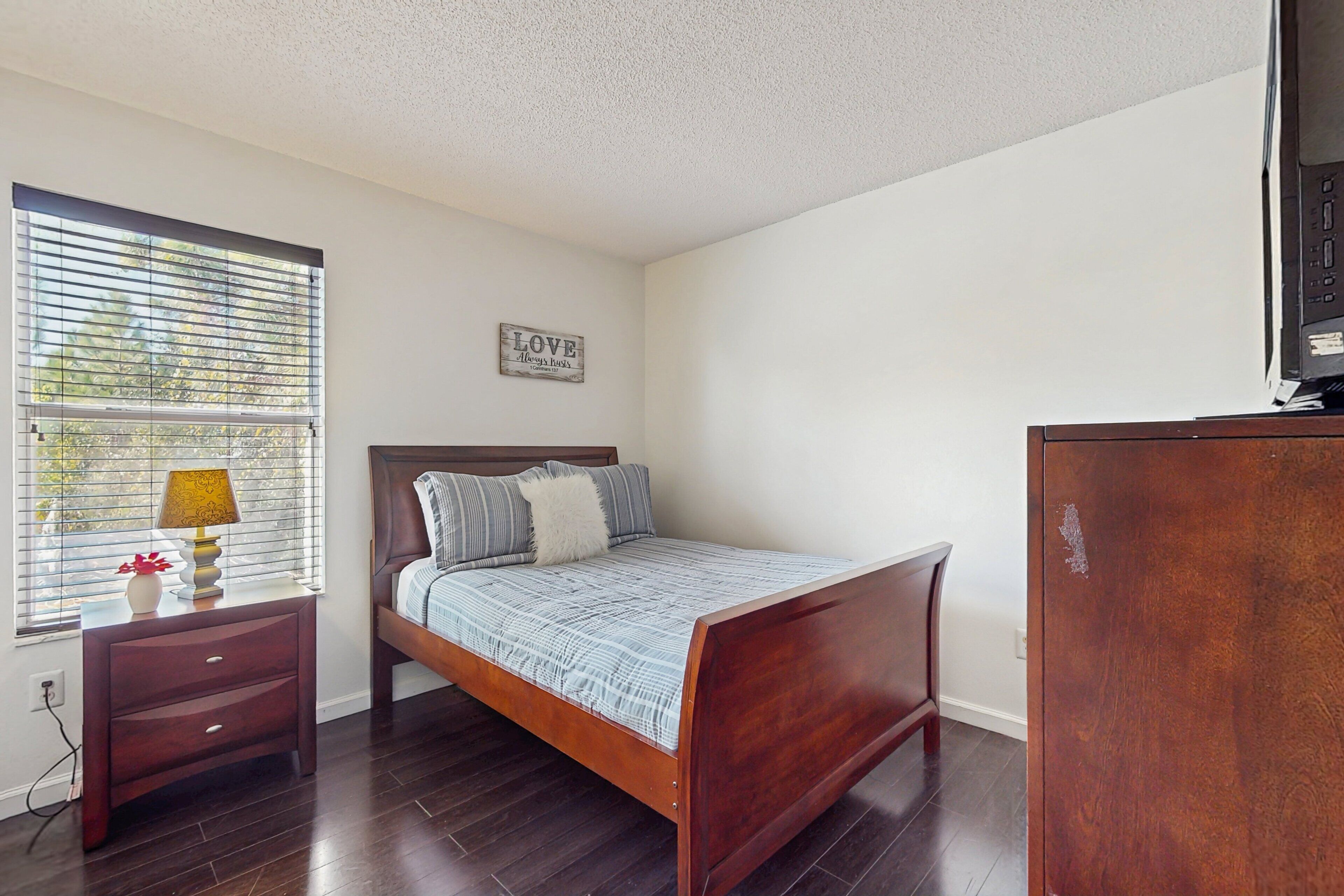 Un dormitorio con una cama de madera, una mesita de noche, una lámpara, un aparador y una ventana con persianas.