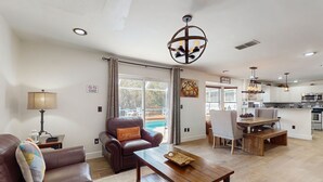Un vaste salon avec un canapé en cuir marron, une table basse en bois et un coin repas avec un banc en bois. Un lustre est suspendu au plafond, et on aperçoit une cuisine au fond.
