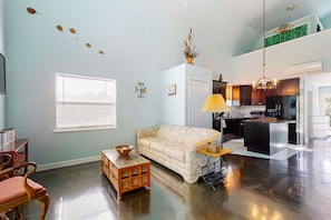 A living room with a sofa, a coffee table, a wooden side table, a window with blinds, a wall-mounted decoration, and a kitchen area with a refrigerator and a hanging light fixture.