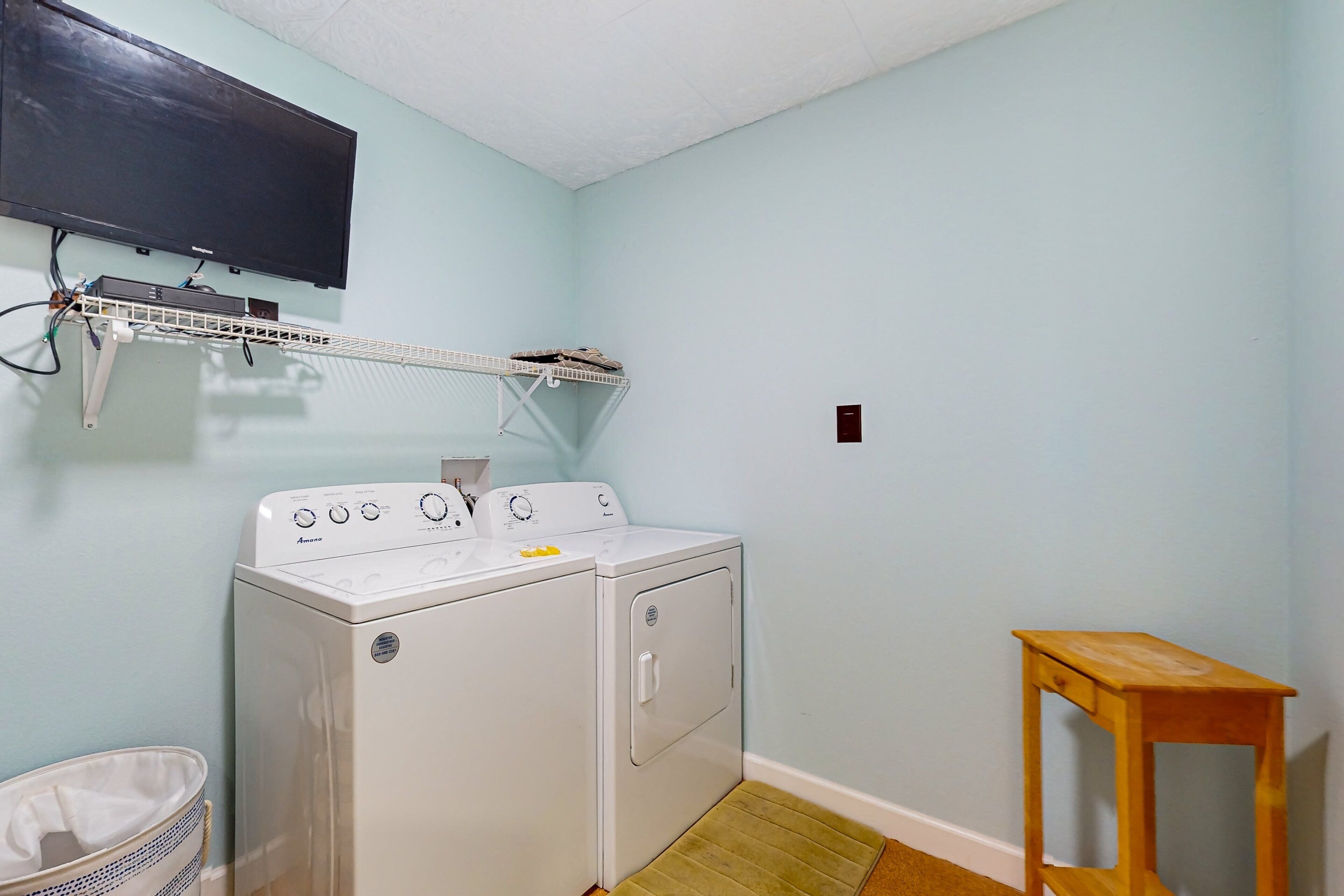 A laundry room with a washing machine, a drying rack, a wooden stool, and a trash bin.