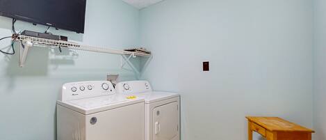 A laundry room with a washing machine, a drying rack, a wooden stool, and a trash bin.