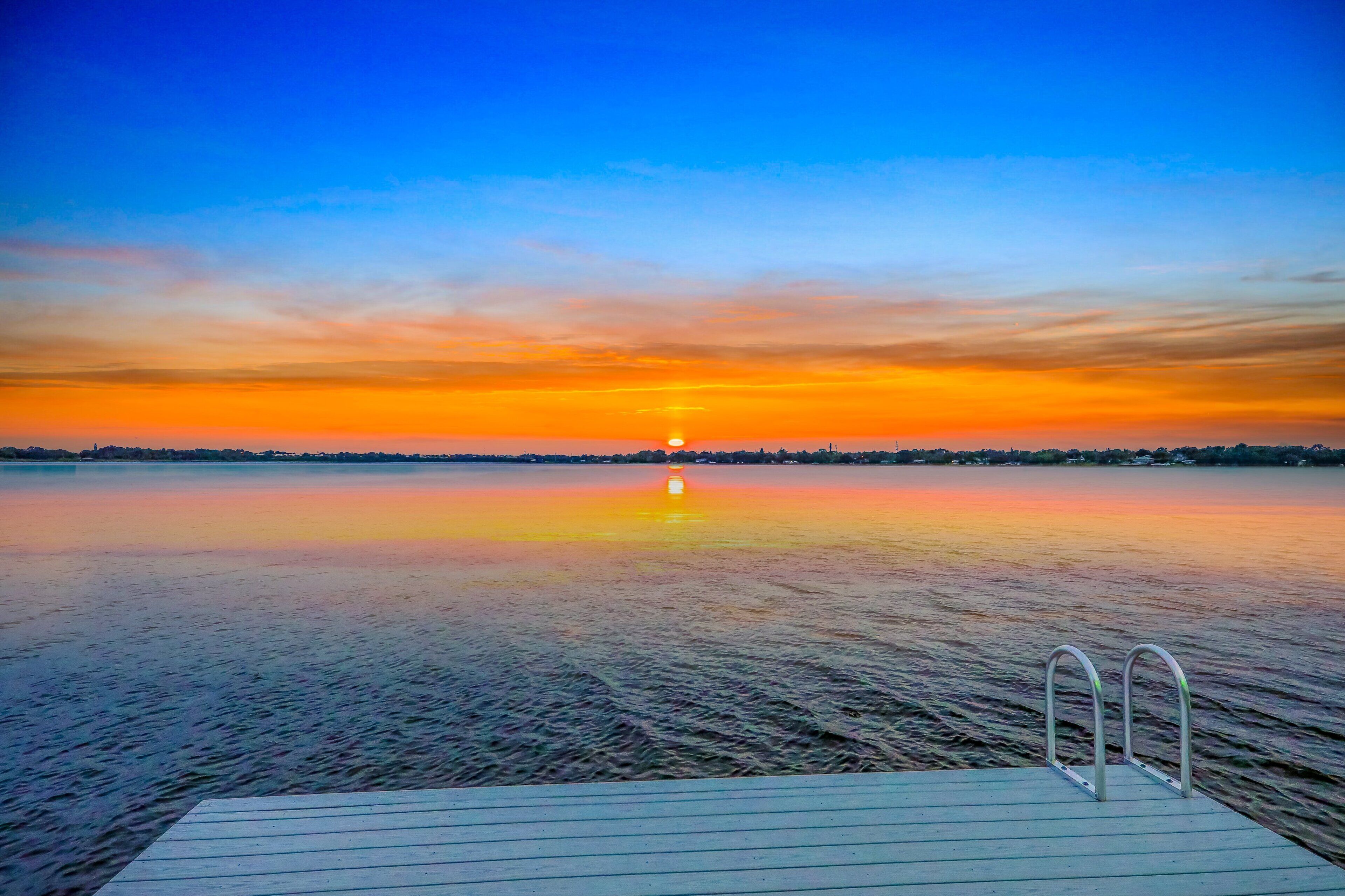 A calm lake at sunset with a wooden dock and metal railings.