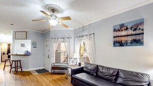 A living room with a black leather sofa, wooden flooring, a ceiling fan, and a painting of a sunset. - Sunny Condo With Pool, hot Tub, Tennis Court, & gym - Balcony & Washer/dryer (Kissimmee)