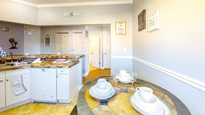 A kitchen with white cabinetry, a dishwasher, a dining table with white dishes, and a hallway with a door. - Sunny Condo With Pool, hot Tub, Tennis Court, & gym - Balcony & Washer/dryer (Kissimmee)