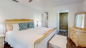 Un dormitorio con cama, ventilador de techo, mesita de noche, cómoda, espejo y puerta que da a un baño.