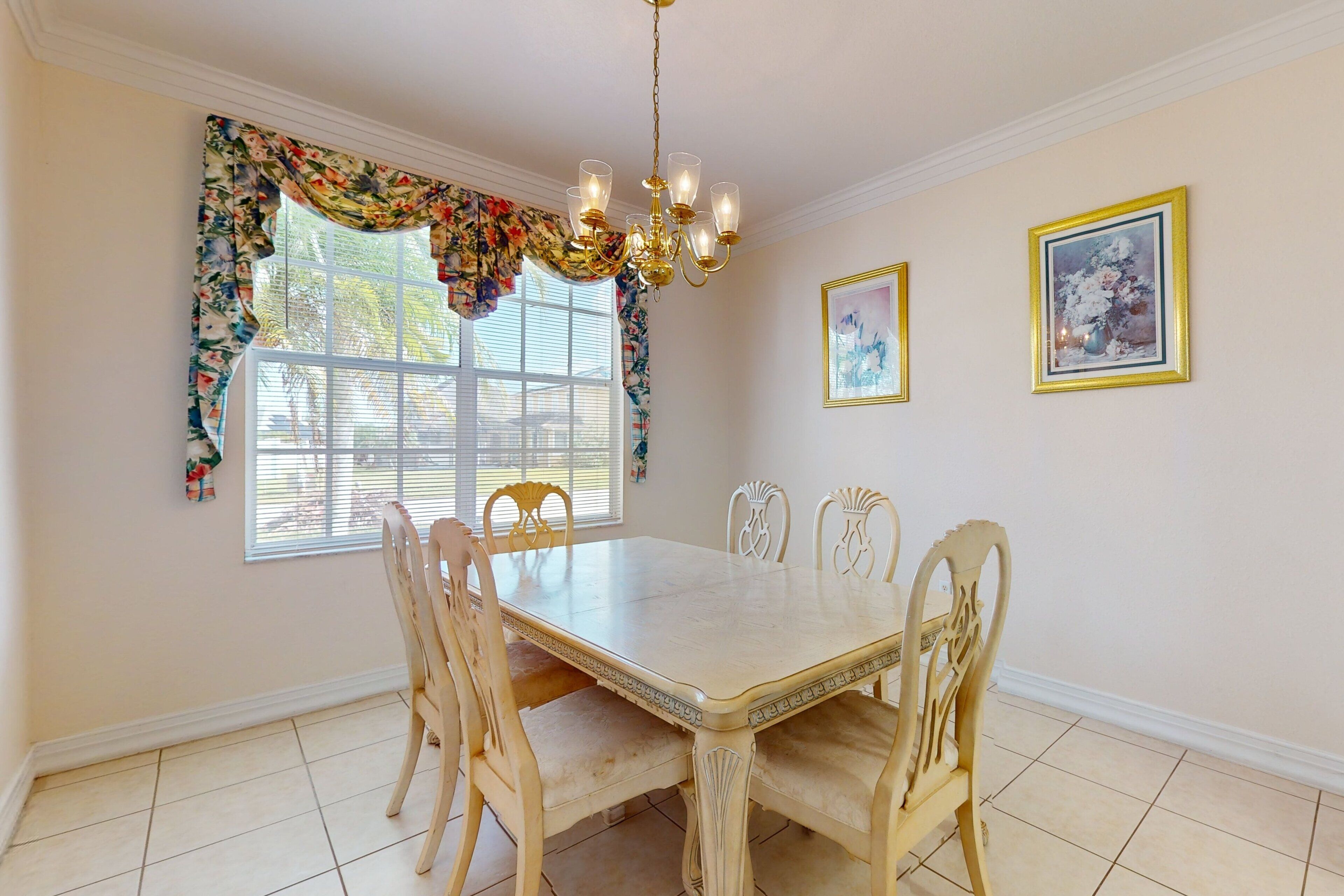 A dining room with a large table, six chairs, a chandelier, floral window valance, and two framed pictures on the wall.