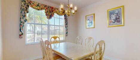 A dining room with a large table, six chairs, a chandelier, floral window valance, and two framed pictures on the wall.