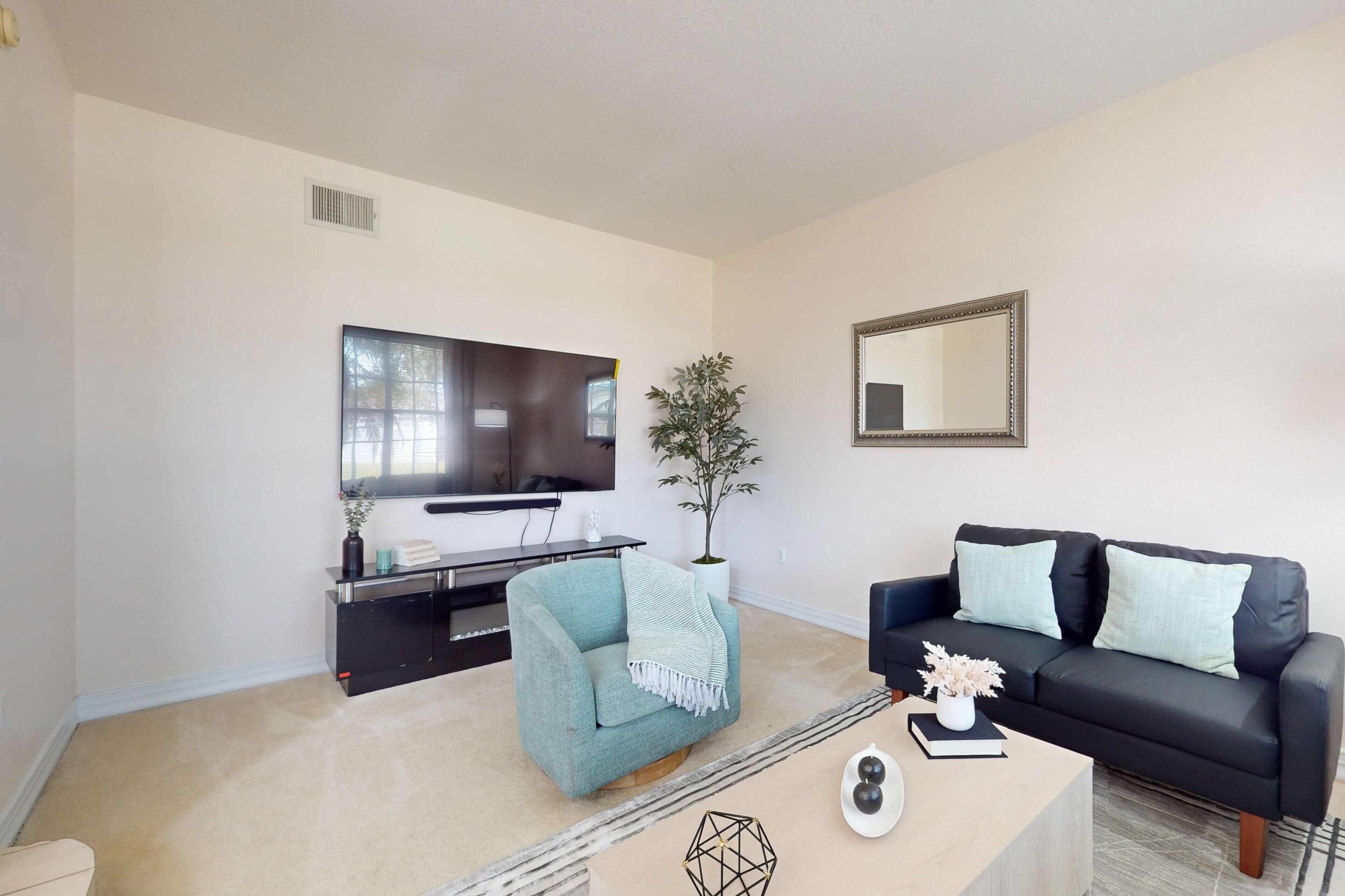 A living room with a flat-screen TV, a black leather sofa, a teal armchair, a coffee table, and a potted plant.