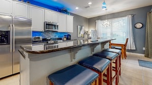 A modern kitchen with a stainless steel refrigerator, microwave, and a built-in oven. There are bar stools along the kitchen island and a dining area with a table and chairs. - All-suite Retreat With Pool, Spa, Waterslides, Tennis - Near Dining & Disney (Kissimmee)