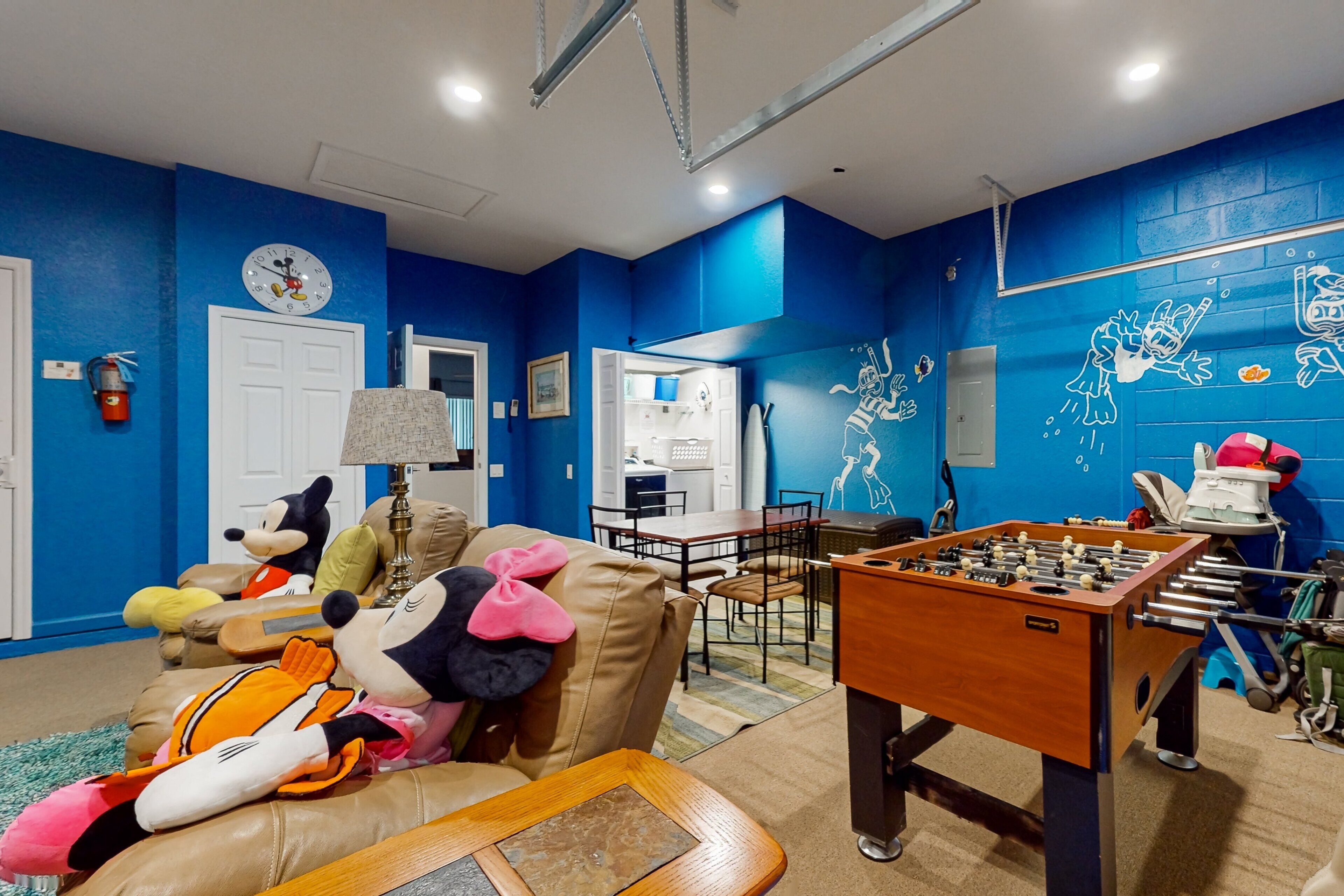 A room with a foosball table, a sofa with plush toys, a dining area, and a blue wall with a clock.