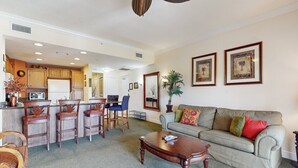 A living room with a sofa, coffee table, and chairs, a kitchen area with cabinets and appliances, and framed artwork on the walls. - Golf Course-front Condo With Golf Views, Balcony, Pool, Fast Wifi, Washer/dryer (Kissimmee)