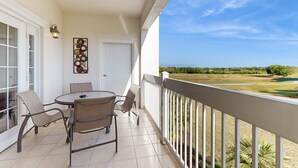 A balcony with a view of a golf course, a dining table with chairs, and a wall-mounted light fixture. - Golf Course-front Condo With Golf Views, Balcony, Pool, Fast Wifi, Washer/dryer (Kissimmee)