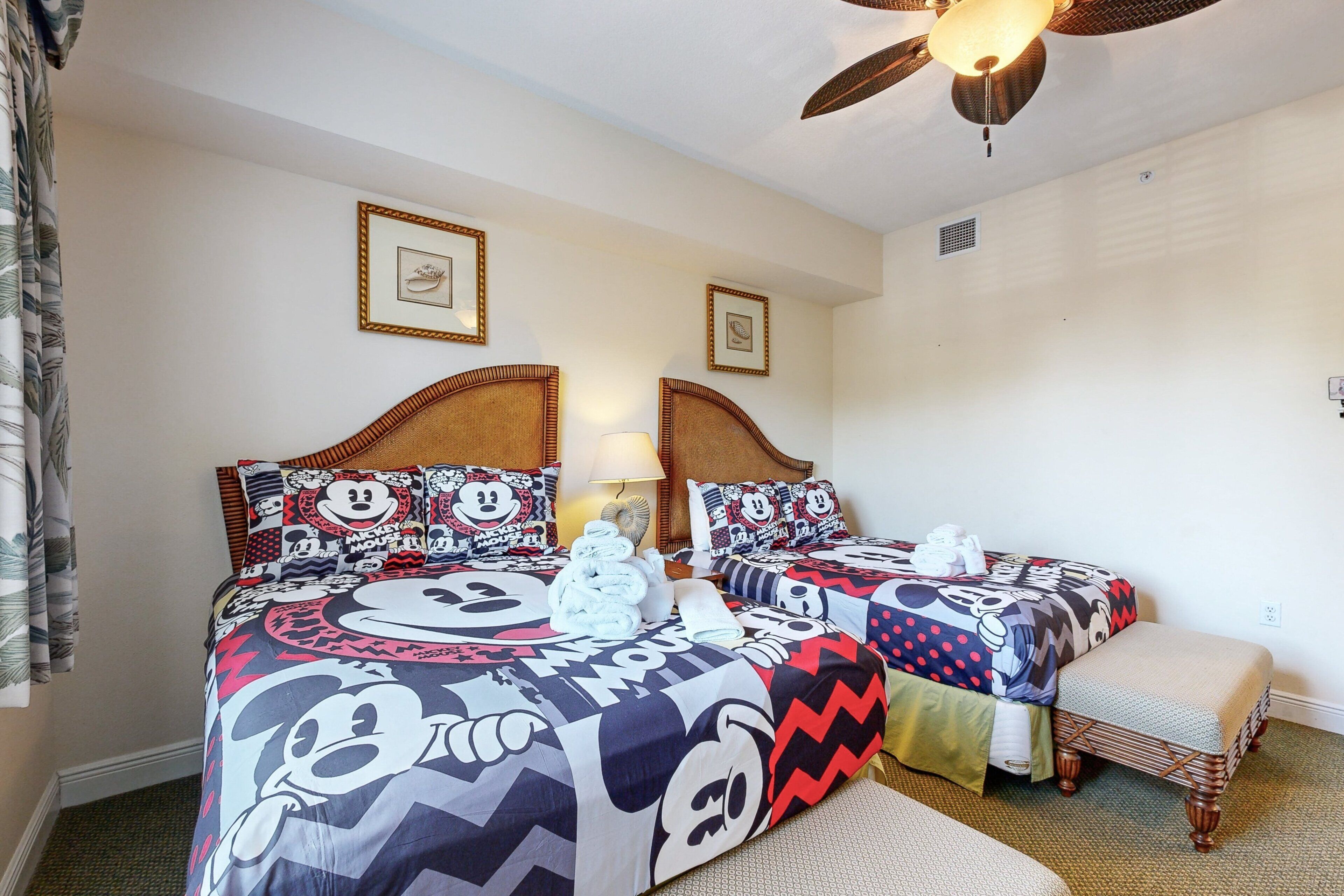 Habitación de hotel con dos camas, ambas con ropa de cama temática de Mickey Mouse, un ventilador de techo y una mesita con lámpara.