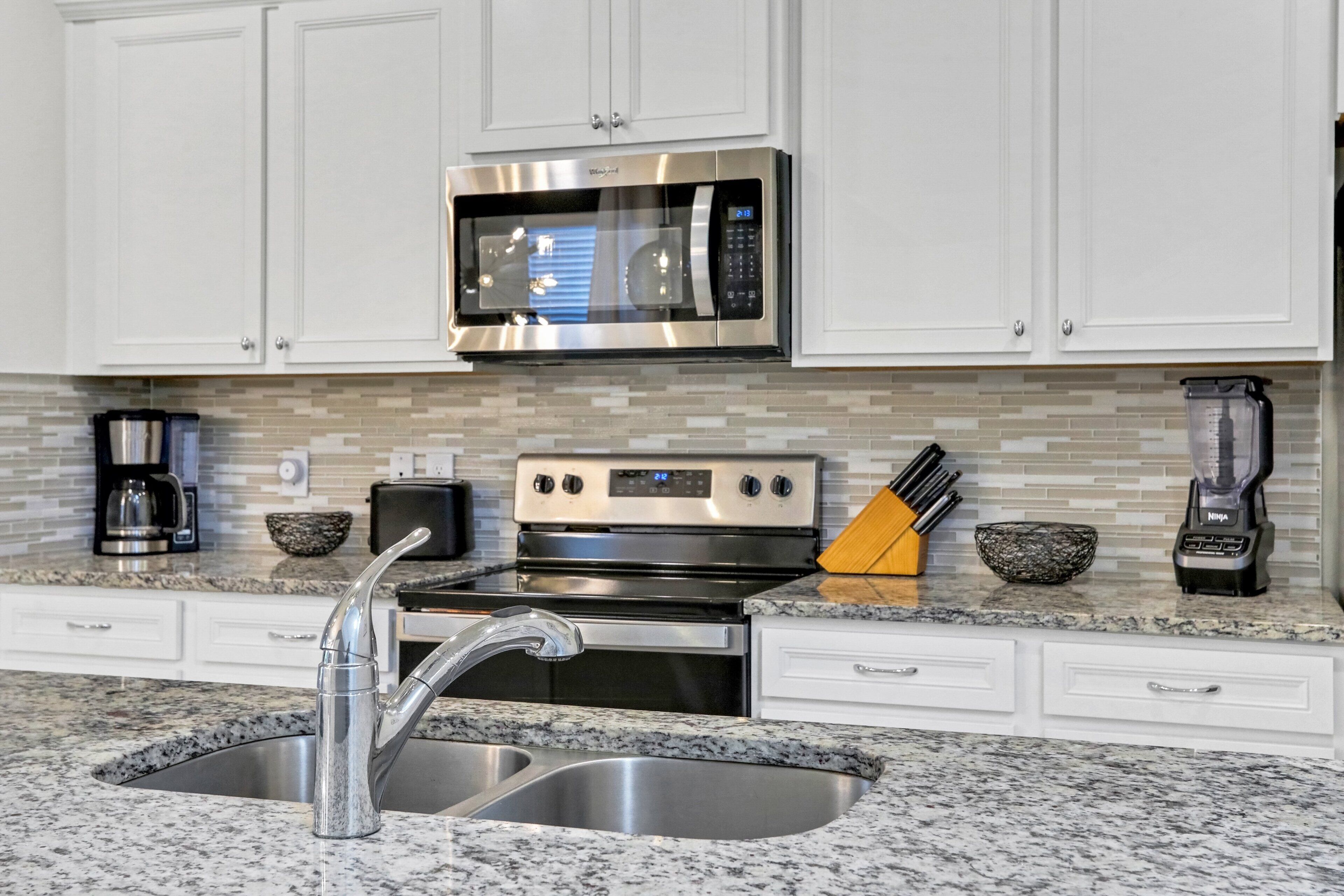 A kitchen with white cabinets, a stainless steel microwave, a stove, a sink, and a coffee maker.