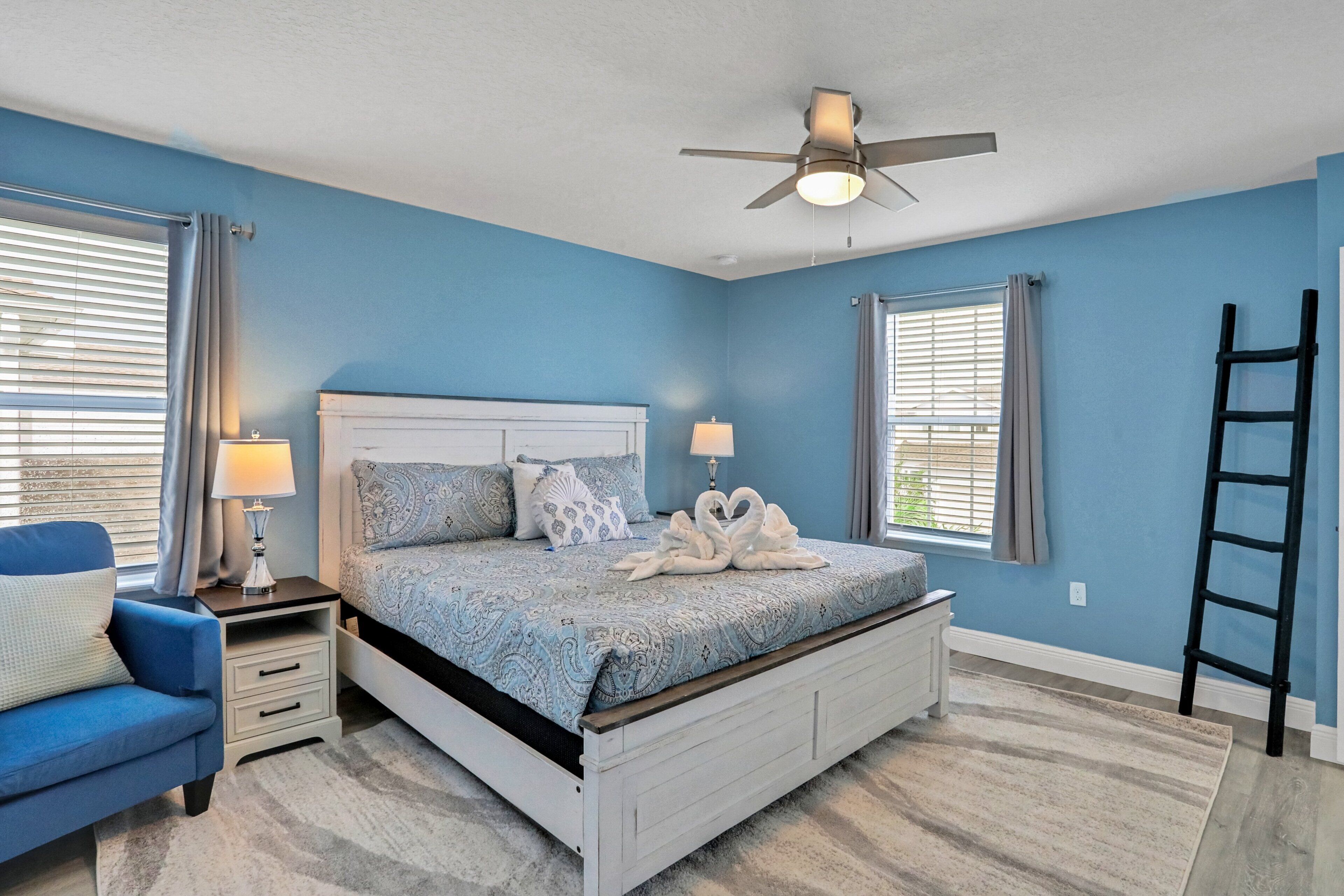 A bedroom with a bed, a nightstand, a lamp, a chair, a ceiling fan, and a window with blinds.