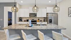 A modern kitchen with granite countertops, white cabinetry, a stainless steel refrigerator, and a central island with bar stools. - Relax & Play Resort Villa Magical 6-bedroom Retreat Near Disney! (Davenport)
