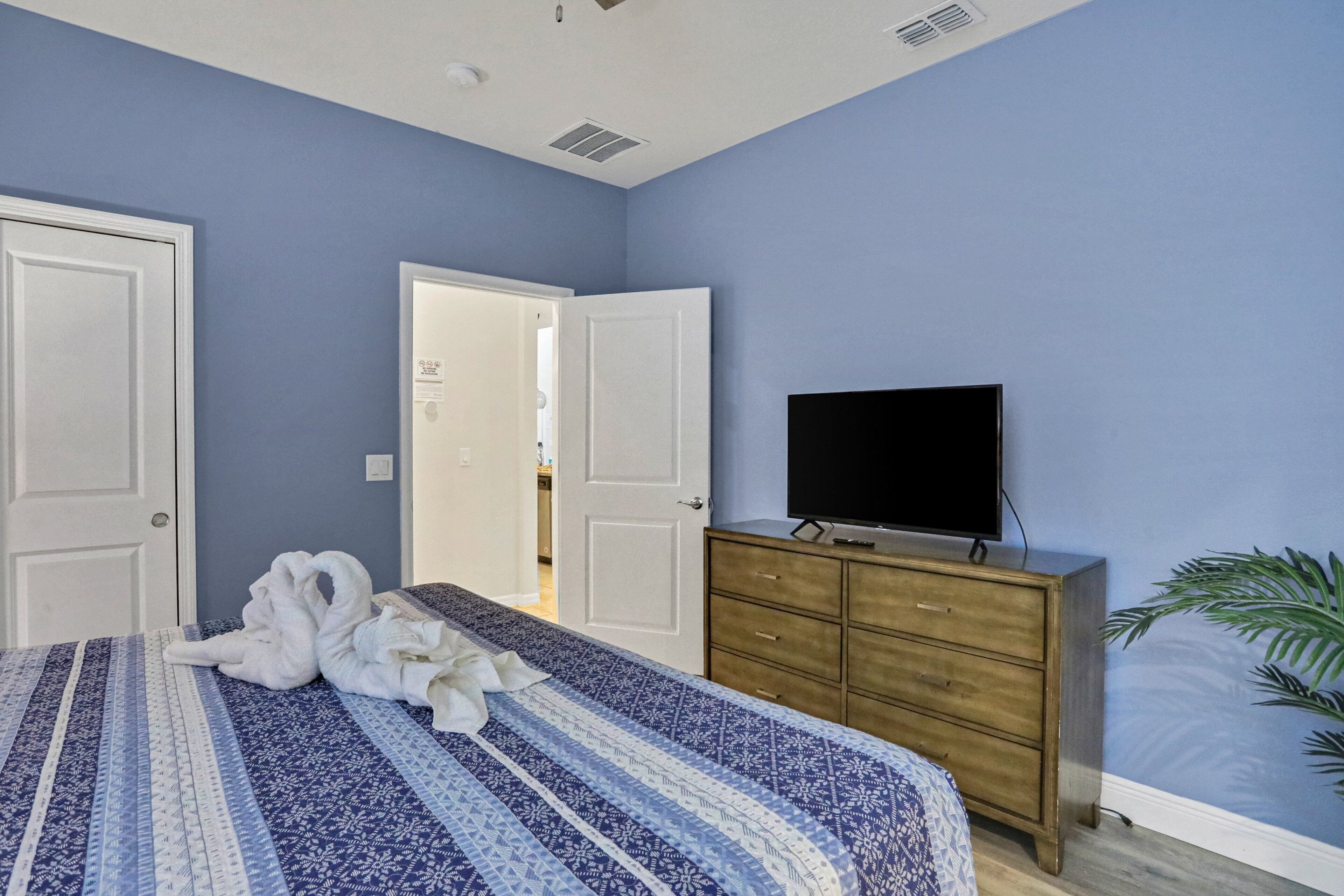Une chambre à coucher avec un lit, un téléviseur posé sur une commode en bois, et une porte donnant sur une autre pièce.