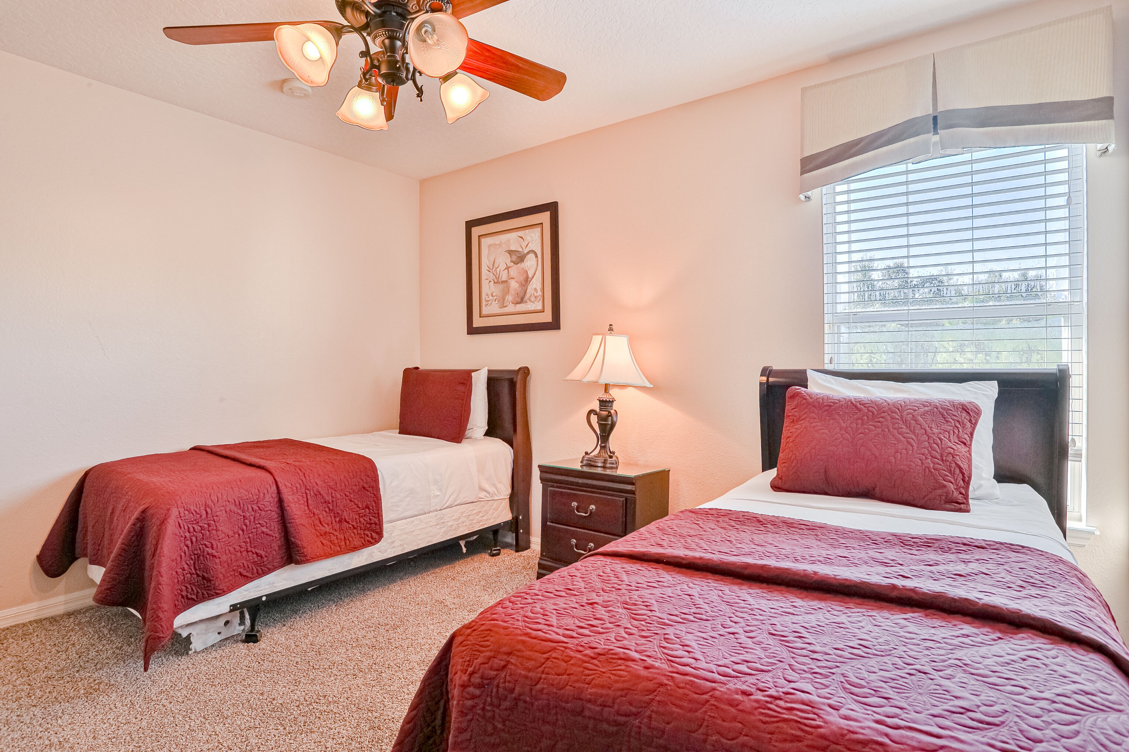 A bedroom with two beds, a ceiling fan, a nightstand with a lamp, and a window with blinds.