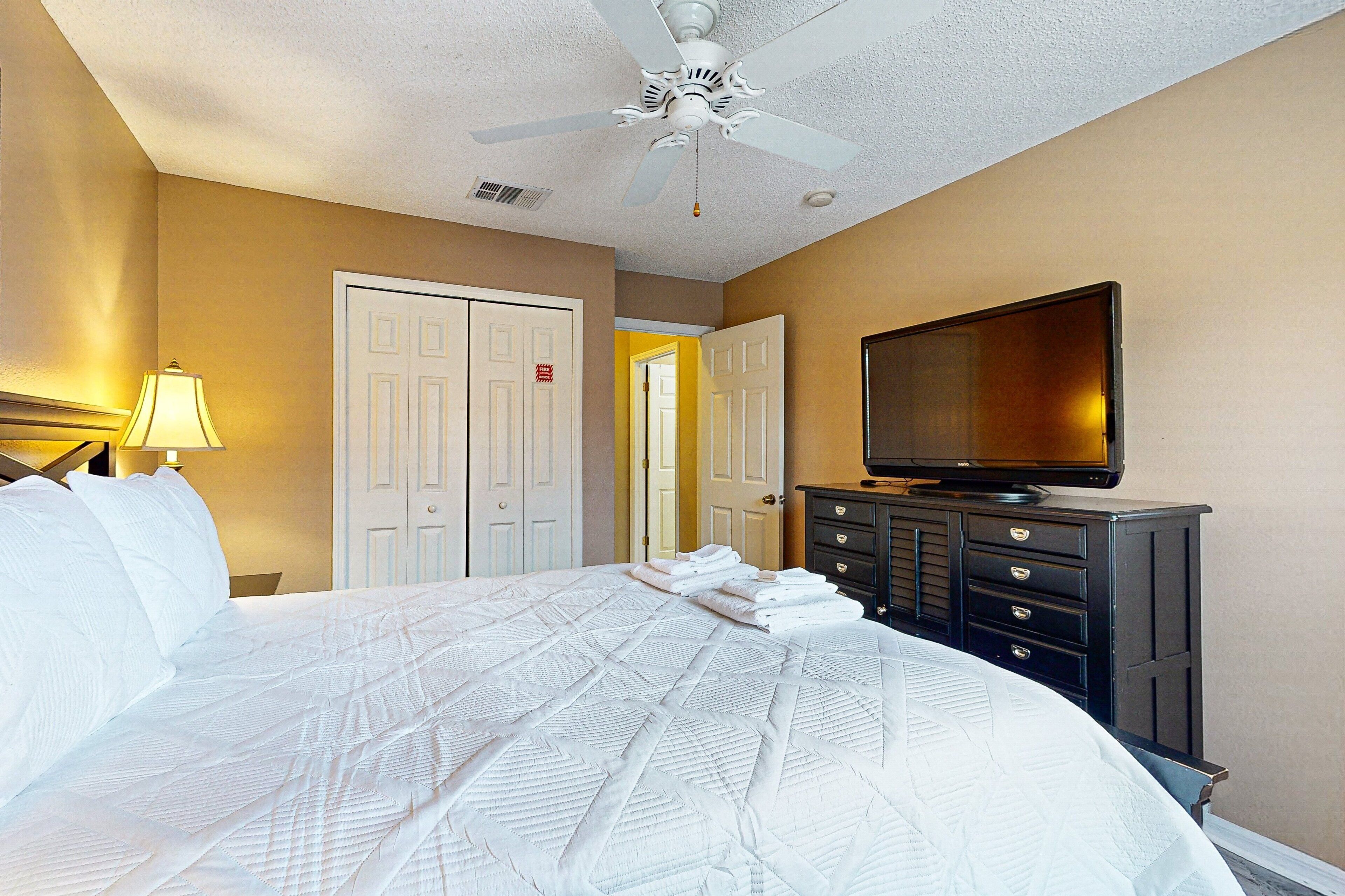 A hotel room with a bed, a television on a dresser, a ceiling fan, and a door leading to another room.