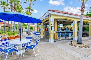 Uma área de pátio ao ar livre com cadeiras e mesas listradas em azul e branco, um bar com balcão e banquetas, e palmeiras ao fundo.