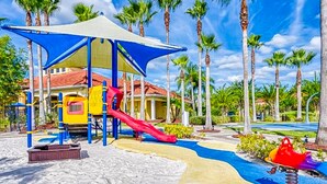A playground with a slide and swings, surrounded by palm trees and a building with a red roof. - Beautiful & Spacious Resort Home in the Heart of Central FL (Kissimmee)