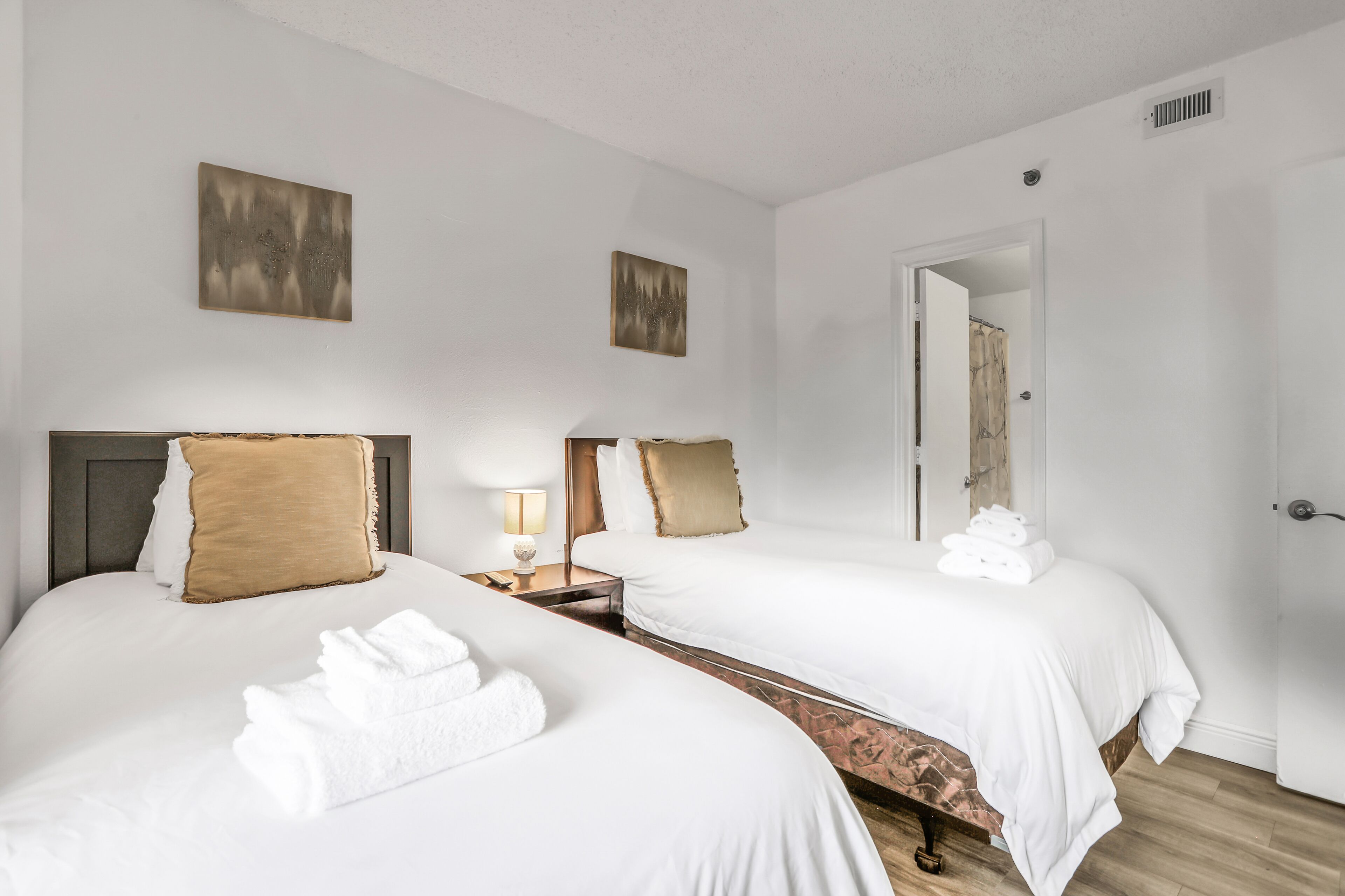 Una habitación de hotel con dos camas, ropa de cama blanca, un cabecero de madera, una mesita de noche con lámpara y obras de arte en las paredes.