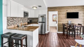 A modern kitchen with white cabinets, a stainless steel refrigerator, a microwave, and a tiled backsplash. There is a wooden dining area with a table and chairs.