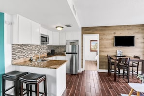 Eine moderne Küche mit weißen Schränken, einem Edelstahl-Kühlschrank, einer Mikrowelle und einer gefliesten Spritzschutzwand. Es gibt einen Essbereich mit Holztisch und Stühlen.