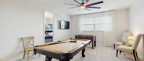 A pool table, a foosball table, a ceiling fan, a TV, and a window with curtains.