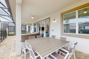 A covered patio area with a wooden table and white chairs, a ceiling fan, and a view of the interior through a glass door. - Solara Joy Disney-themed Retreat in Kissimmee With Stellar Resort Amenities (Kissimmee)