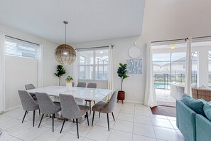 A modern dining area with a white table and grey chairs, large windows with white curtains, and a view of a pool outside. - Solara Joy Disney-themed Retreat in Kissimmee With Stellar Resort Amenities (Kissimmee)