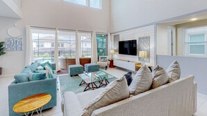 A spacious living room with a high ceiling, large windows, a flat-screen TV, and a comfortable seating area with sofas and a coffee table. - Solara Joy Disney-themed Retreat in Kissimmee With Stellar Resort Amenities (Kissimmee)