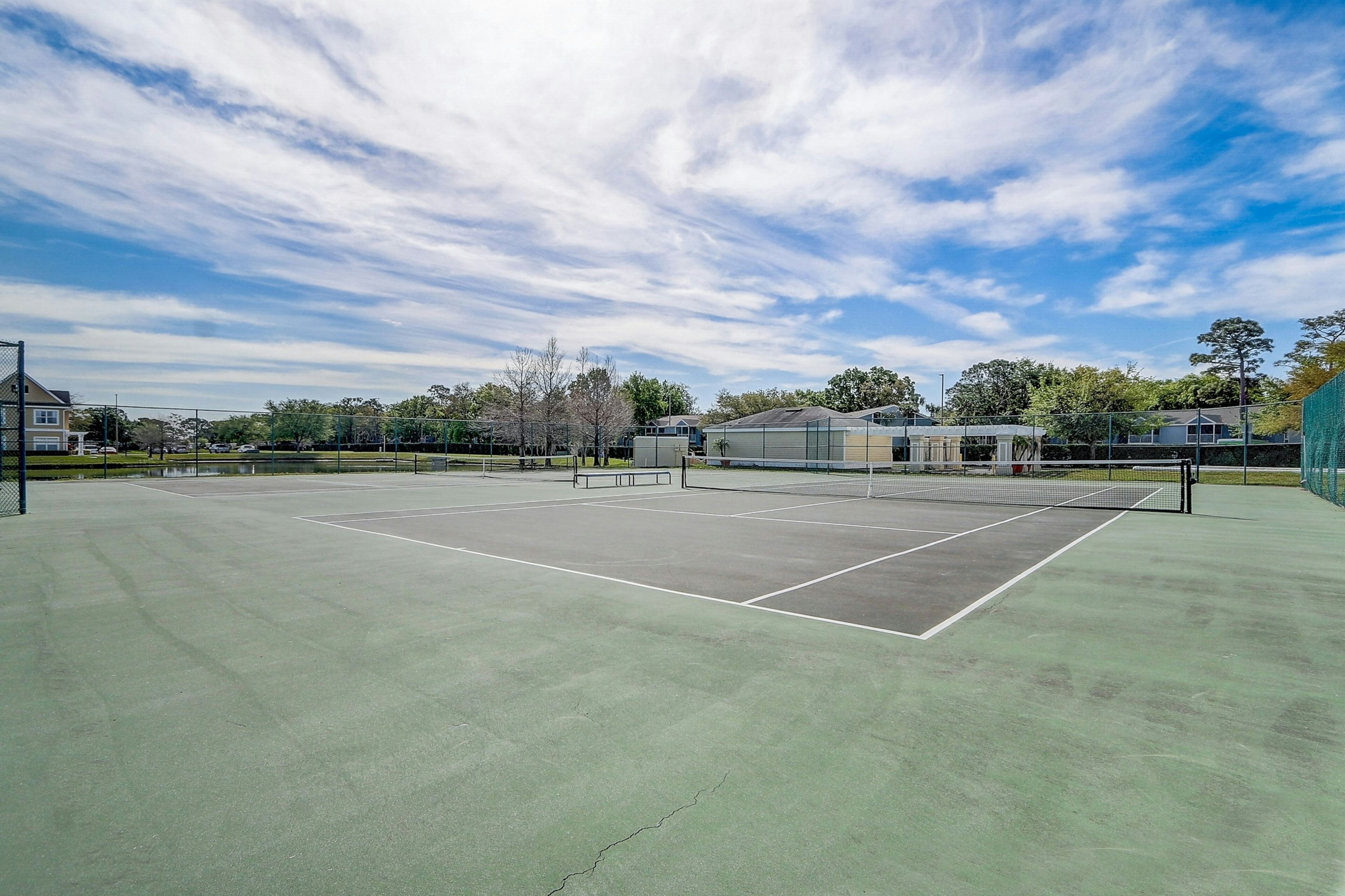 Un court de tennis en plein air, doté d’un filet, entouré d’arbres et de bâtiments.