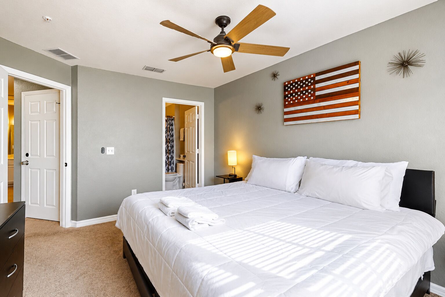 Une chambre d’hôtel avec un lit, un ventilateur de plafond, un drapeau américain accroché au mur et une salle de bain visible à travers une porte ouverte.