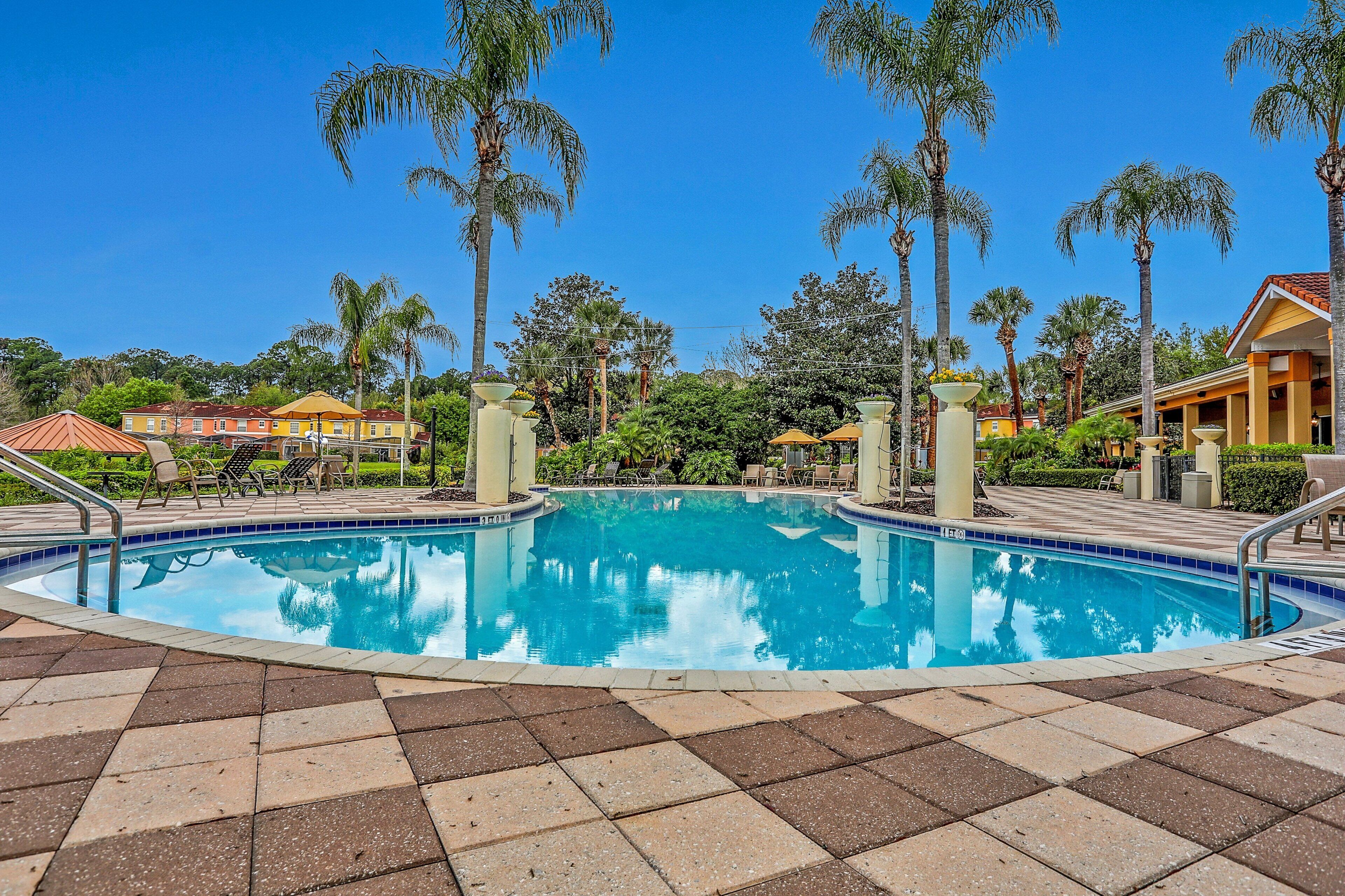 A swimming pool with a tiled deck, surrounded by palm trees and a building with a red roof.