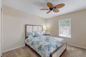 Ein Schlafzimmer mit einem Bett, einem Deckenventilator, einem Nachttisch, einer Lampe und einem Fenster.