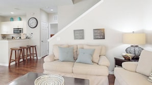A living room with a sofa, a coffee table, and a lamp. There is a kitchen in the background with a refrigerator, microwave, and stools. - Spacious Family Friendly Home With Resort Style Shared Pool (Kissimmee)