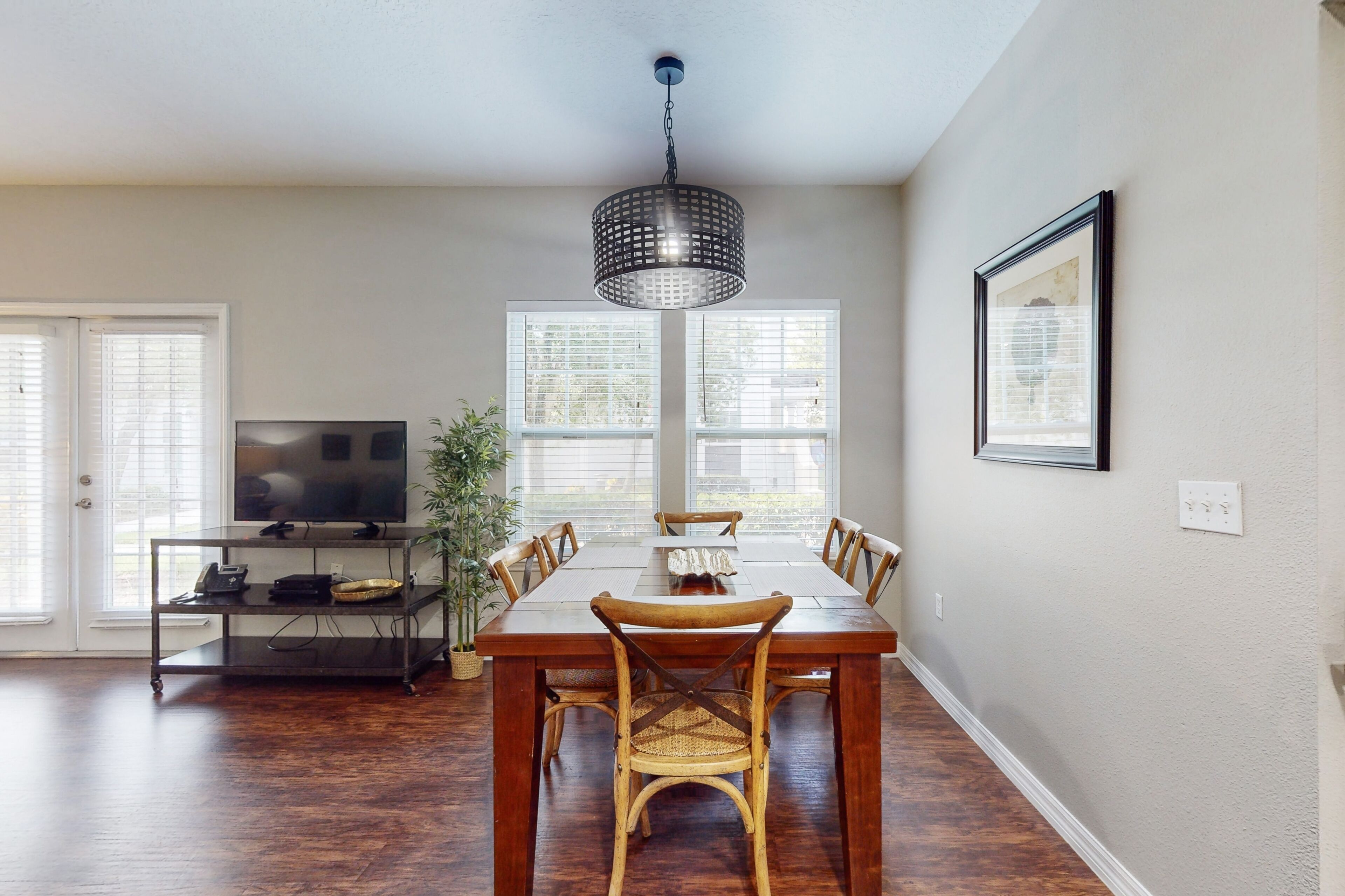 A dining area with a wooden table, six chairs, a TV on a stand, and a hanging light fixture.
