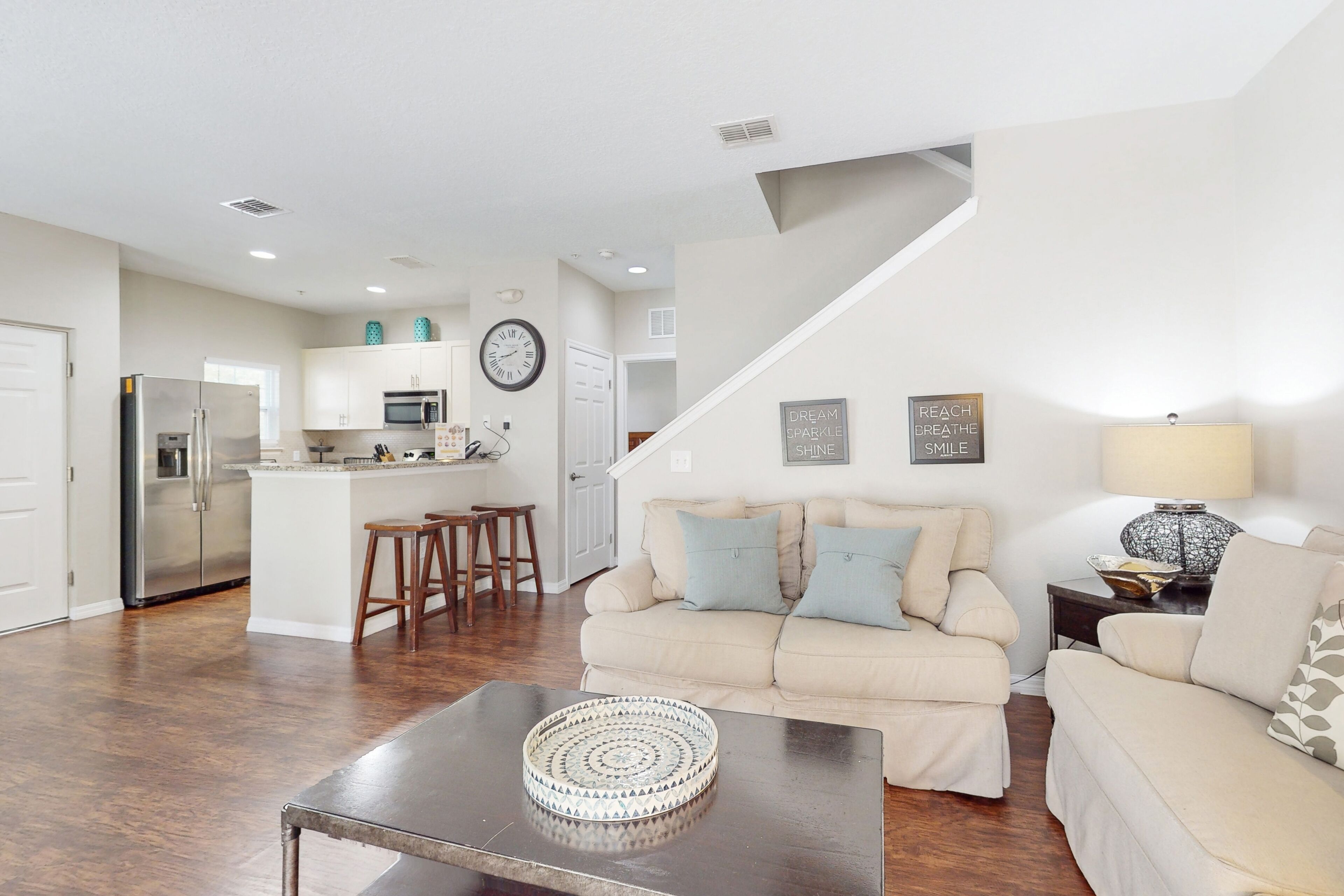 A modern kitchen with stainless steel appliances, a dining area with a round coffee table, and a living room with a beige sofa and decorative pillows.