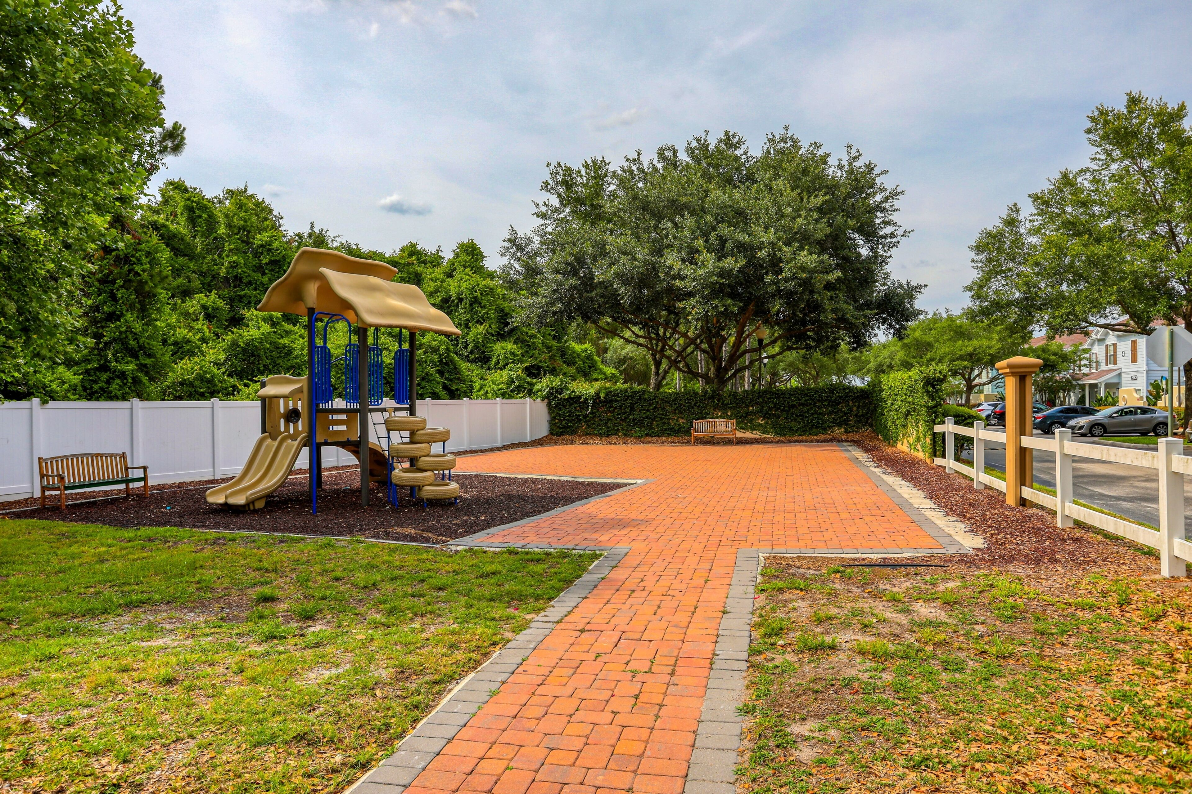 A playground with a slide and climbing structures, surrounded by a brick pathway and greenery.