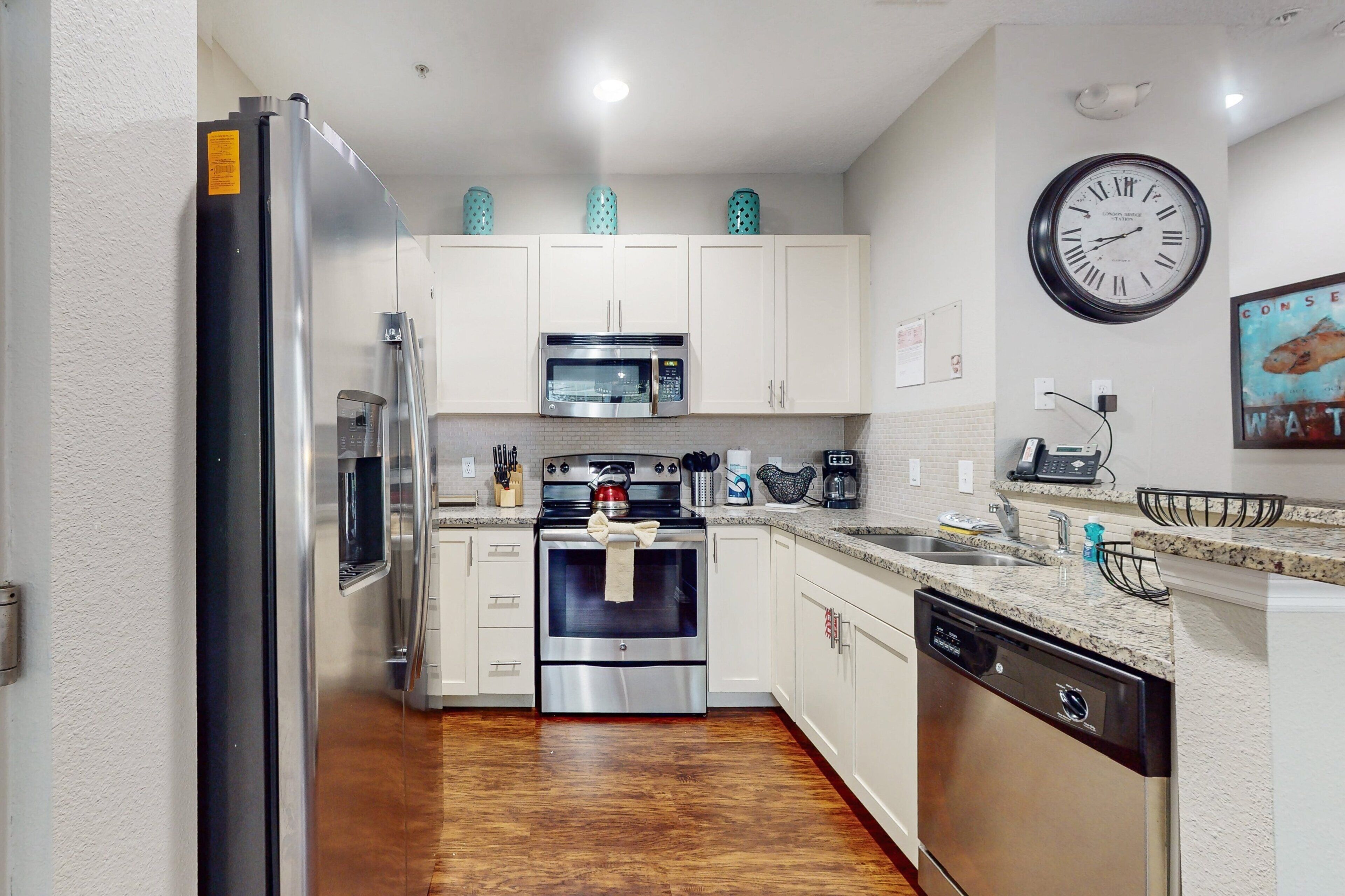 A modern kitchen with stainless steel appliances, white cabinets, a wooden floor, and a wall clock.
